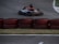 A go-kart zooms around a racetrack, with a driver wearing a helmet and racing suit. Red and black tires are stacked as barriers along the track. The asphalt is dark, and the grass is visible along the edge.