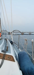 A sailing boat is navigating on a calm body of water. The deck displays ropes, a wooden railing, and cushioned blue pads. In the background, a stone bridge with repeating arches spans across the water, while the sky is clear and slightly overcast.