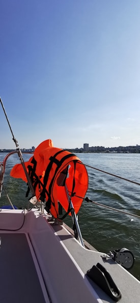 A bright orange life jacket is hanging on the railing of a boat, with a view of calm water and a distant city skyline under a clear blue sky.