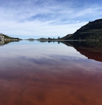 A tranquil lake reflects the sky and surrounding hills. The water appears reddish, likely due to sediment or dissolved minerals. The sky is partly cloudy, and the landscape features gentle slopes and sparse vegetation.