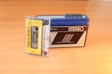 A vintage cassette tape labeled 'Modern Arapça' is partially inserted into a retro portable stereo cassette player placed on a wooden surface. The cassette player has a blue top and a black front with various buttons.