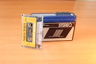 A vintage cassette tape labeled 'Modern Arapça' is partially inserted into a retro portable stereo cassette player placed on a wooden surface. The cassette player has a blue top and a black front with various buttons.