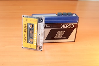 A vintage cassette tape labeled 'Modern Arapça' is partially inserted into a retro portable stereo cassette player placed on a wooden surface. The cassette player has a blue top and a black front with various buttons.