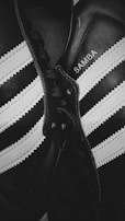 Close-up shot of a pair of classic black and white athletic shoes with the word 'SAMBA' written on one. The shoes feature distinctive white stripes and black laces on a leather surface, highlighting their iconic design.