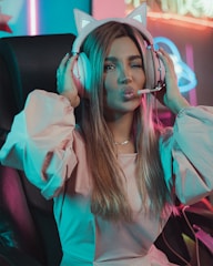A person sitting on a gaming chair wearing cat-ear headphones and making a playful facial expression with a wink and puckered lips. The room is illuminated with neon lights providing a colorful ambiance.