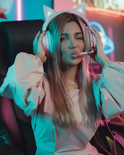 A person sitting on a gaming chair wearing cat-ear headphones and making a playful facial expression with a wink and puckered lips. The room is illuminated with neon lights providing a colorful ambiance.