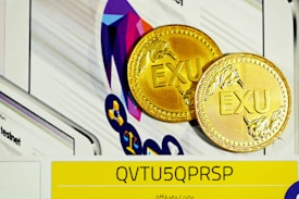 Two gold coins with the letters 'EXU' embossed on them are placed against a colorful, abstract geometric background. In the foreground, a yellow strip with the code 'QVTU5QPRSP' is visible, possibly indicating an affiliate or promotional code.
