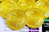 Several gold coins are displayed, each with the letters 'EXU' embossed on them. The coins feature intricate designs and are arranged on top of a document or screen displaying numerical data and text.