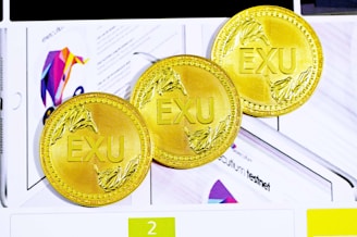 Three gold coins with the letters EXU embossed in the center. Each coin features intricate leaf designs on the edges. They are placed against a background that includes colorful graphics and partially visible text.