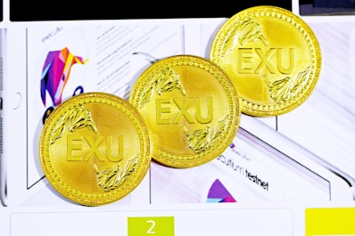 Three gold coins with the letters EXU embossed in the center. Each coin features intricate leaf designs on the edges. They are placed against a background that includes colorful graphics and partially visible text.