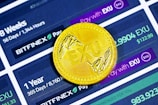 A golden coin with the letters 'EXU' embossed on it is placed on a digital interface displaying cryptocurrency exchange information. The interface shows details like '56 Days / 1,344 Hours', '365 Days / 8,760 Hours', 'BITFINEX', and different payment amounts denoted in 'EXU'.