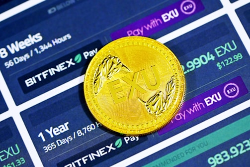 A golden coin with the letters 'EXU' embossed on it is placed on a digital interface displaying cryptocurrency exchange information. The interface shows details like '56 Days / 1,344 Hours', '365 Days / 8,760 Hours', 'BITFINEX', and different payment amounts denoted in 'EXU'.