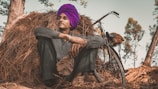 A person wearing a vibrant purple turban is sitting in a relaxed posture against a stack of hay. The individual is dressed in casual attire and is beside a bicycle. The background includes trees with sparse green leaves, suggesting a rural setting.