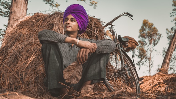 A person wearing a vibrant purple turban is sitting in a relaxed posture against a stack of hay. The individual is dressed in casual attire and is beside a bicycle. The background includes trees with sparse green leaves, suggesting a rural setting.
