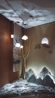 A serene indoor setup features an artistic wall design with subtle lighting. A tree branch extends toward the ceiling, surrounded by decorative clouds and a moon-like light source. The floor area presents an elaborate Zen garden with rocks and sand art, adding to the tranquil ambiance.