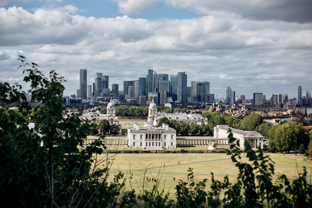 green grass field near city buildings under white clouds and blue sky during daytime