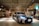 A sleek, silver luxury car is parked in a modern, well-lit indoor parking area. The ceiling features stylish wooden slats with spotlights, adding a sophisticated ambiance. A small arrangement of flowers is visible to the right, near a polished wall.
