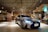 A sleek, silver luxury car is parked in a modern, well-lit indoor parking area. The ceiling features stylish wooden slats with spotlights, adding a sophisticated ambiance. A small arrangement of flowers is visible to the right, near a polished wall.