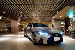 A sleek, silver luxury car is parked in a modern, well-lit indoor parking area. The ceiling features stylish wooden slats with spotlights, adding a sophisticated ambiance. A small arrangement of flowers is visible to the right, near a polished wall.