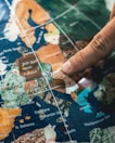 A close-up shot of a person's finger pointing at a specific location on a colorful map. The map includes regions of Europe with labels for cities and countries. The focus is on the Mediterranean area, including Italy, France, and Greece.