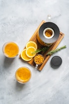A flat lay of a wooden tray with a glass teapot containing a golden liquid. Sliced oranges and a measuring cup of dried berries are next to the teapot. Two glasses filled with the same golden liquid are placed nearby. A sprig of fresh rosemary adds a touch of green.