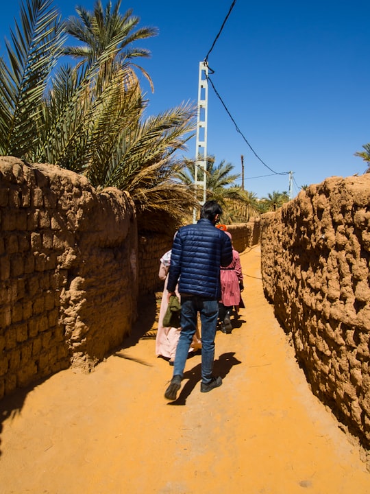 man in red shirt and blue denim jeans walking on brown dirt road during daytime in Béni Abbès Algeria