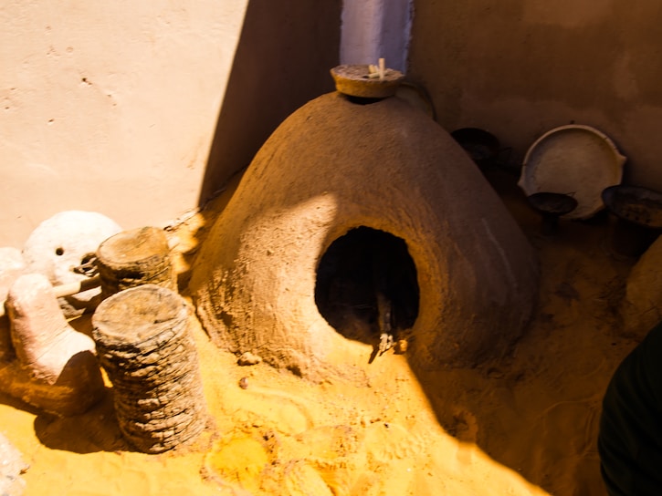 A traditional clay oven is situated outdoors with several metal and stone objects nearby. The structure of the oven has a dome shape, blending into the sandy environment. Nearby, there are small stacks of what appear to be circular stones, and various metal plates are leaning against the wall.