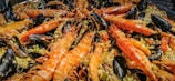 A close-up view of a seafood dish featuring large prawns, mussels, and rice. The prawns are prominently displayed in the center, with their vibrant orange color contrasting against the dark shells of the mussels. The rice is mixed with vegetables, adding texture and color to the dish.