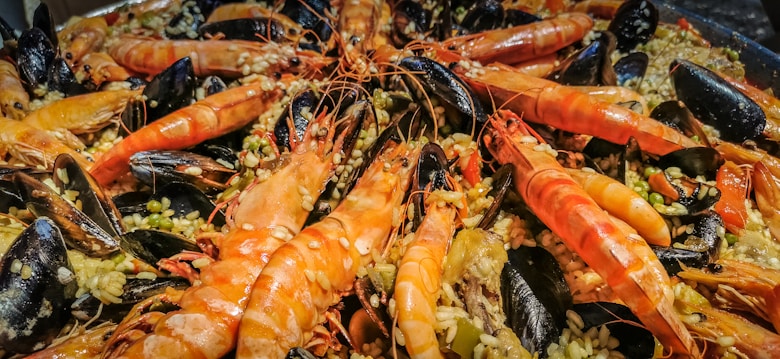A close-up view of a seafood dish featuring large prawns, mussels, and rice. The prawns are prominently displayed in the center, with their vibrant orange color contrasting against the dark shells of the mussels. The rice is mixed with vegetables, adding texture and color to the dish.