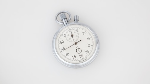 A vintage analog stopwatch with a metallic case and a white dial featuring black numbers. The watch has two dials; the main dial is marked with seconds while the smaller one is for minutes. The top has a winding crown and push button.