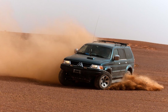 A rugged SUV is driving across a desert landscape, kicking up a large cloud of dust. The terrain is dry and sandy, creating a sense of motion and adventure as the vehicle maneuvers through the open space.