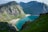 A breathtaking view of a secluded beach cove surrounded by towering, rugged mountains. The mountains are covered with patches of lush greenery, and the water in the cove is a vibrant turquoise blue. The scene is serene and isolated, with a sense of untouched natural beauty.