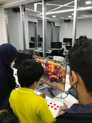 A group of children is gathered around a desktop computer, engaged in playing a colorful, action-packed arcade game displayed on the monitor. The setting appears to be a modern, well-lit office or gaming space with large glass windows, showing multiple computer workstations in the background.