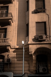 A vintage building facade with multiple windows, balconies, and air conditioning units. The building is made of a light-colored stone, and the image captures the play of light and shadow on its textured surface. A streetlight is installed in front of the building, providing a modern contrast to the older architecture.