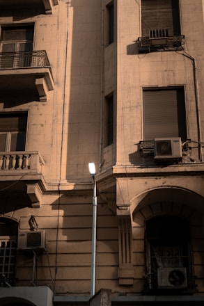 A vintage building facade with multiple windows, balconies, and air conditioning units. The building is made of a light-colored stone, and the image captures the play of light and shadow on its textured surface. A streetlight is installed in front of the building, providing a modern contrast to the older architecture.