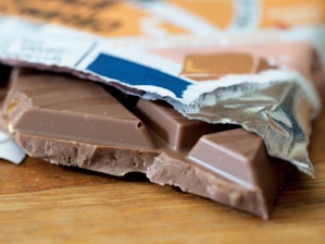 A partially unwrapped chocolate bar rests on a wooden surface. The bar is broken, revealing smooth milk chocolate and an intricate design on its surface. The wrapper, with a mix of colors including blue, orange, and white, is crumpled around the chocolate.
