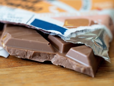 A partially unwrapped chocolate bar rests on a wooden surface. The bar is broken, revealing smooth milk chocolate and an intricate design on its surface. The wrapper, with a mix of colors including blue, orange, and white, is crumpled around the chocolate.
