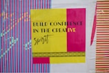A bold and colorful poster features the phrase 'Build Confidence in the Creative Spirit' in black and yellow text. The background displays vibrant vertical stripes in blue and pink, with contrasting designs in yellow and purple.