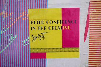 A bold and colorful poster features the phrase 'Build Confidence in the Creative Spirit' in black and yellow text. The background displays vibrant vertical stripes in blue and pink, with contrasting designs in yellow and purple.