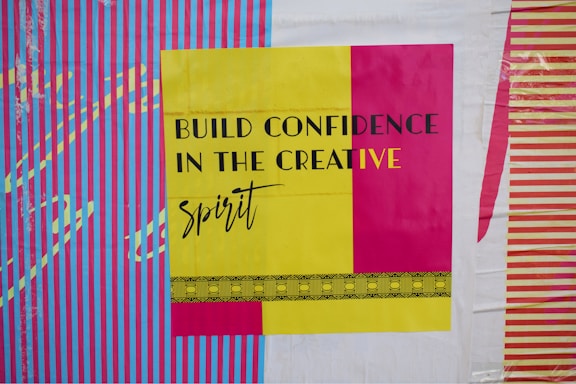 A bold and colorful poster features the phrase 'Build Confidence in the Creative Spirit' in black and yellow text. The background displays vibrant vertical stripes in blue and pink, with contrasting designs in yellow and purple.