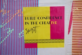 A bold and colorful poster features the phrase 'Build Confidence in the Creative Spirit' in black and yellow text. The background displays vibrant vertical stripes in blue and pink, with contrasting designs in yellow and purple.
