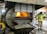 A person in a high-visibility jacket is observing a large industrial furnace that is emitting a bright orange glow. The furnace is situated in an industrial setting with metal structures surrounding it. Ash or debris is visible in a container beneath the furnace.