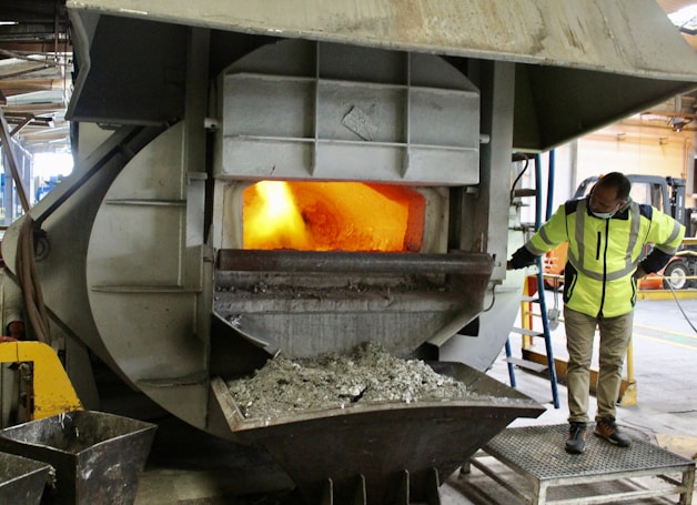 A person in a high-visibility jacket is observing a large industrial furnace that is emitting a bright orange glow. The furnace is situated in an industrial setting with metal structures surrounding it. Ash or debris is visible in a container beneath the furnace.