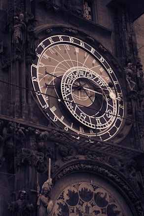 A detailed view of an ornate astronomical clock featuring intricate designs and numerous circular dials with zodiac symbols and Roman numerals. The clock appears to be mounted on an elaborate façade with statues and decorative elements.