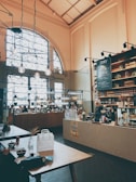 A cozy cafe interior featuring a high ceiling with hanging lights and large arched windows. The space includes a long counter with various items such as drinks and snacks, and a chalkboard menu on the wall. Wooden tables are placed throughout, with patrons sitting and enjoying their meals.