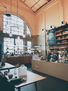 A cozy cafe interior featuring a high ceiling with hanging lights and large arched windows. The space includes a long counter with various items such as drinks and snacks, and a chalkboard menu on the wall. Wooden tables are placed throughout, with patrons sitting and enjoying their meals.