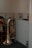 A shiny brass tuba leans against a wall with striped wallpaper. Surrounding it are empty water bottles, both on the floor and lined up on a shelf above. A bouquet of artificial flowers rests on a small table nearby, and the soft lighting creates a calm indoor atmosphere.