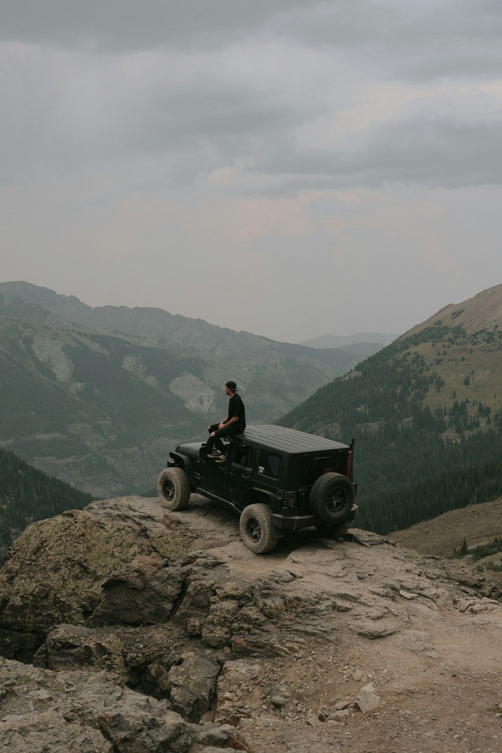 man in black shirt sitting on black jeep wrangler on rocky mountain during daytime