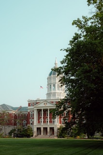 A large, historical building with a distinctive dome and columns, surrounded by lush greenery and an American flag flying in front. The architecture is elegant, featuring red brick and white accents.
