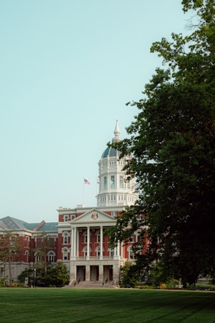 A large, historical building with a distinctive dome and columns, surrounded by lush greenery and an American flag flying in front. The architecture is elegant, featuring red brick and white accents.